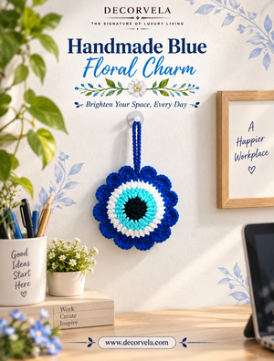 A handmade blue crochet floral charm shaped like an eye, hanging on a wall in a styled home office setting. The image features the brand name 'DECORVELA' and the product title 'Handmade Blue Floral Charm' at the top, with desk props including a plant, a cup, a laptop, and a framed sign.