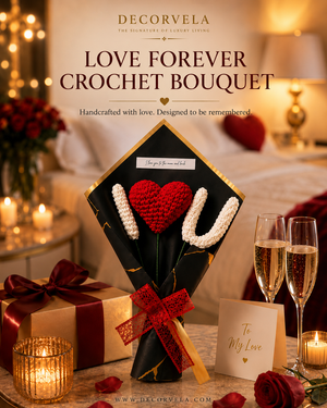 A decorative gift bouquet featuring two crocheted roses with red and white petals wrapped in black paper with gold veining and tied with a red ribbon. The bouquet is displayed on a table with champagne flutes, a gift box, candles, and a card reading 'To My Love' in a warmly lit bedroom setting.