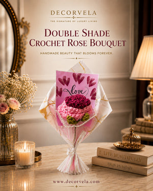 DecorVela Double Shade Crochet Rose Bouquet displayed on a wooden table in a warmly lit, elegant room setting. The bouquet consists of two distinct shades of pink crochet roses wrapped in a white and gold patterned paper sleeve, with a pink 'love' card tucked inside.