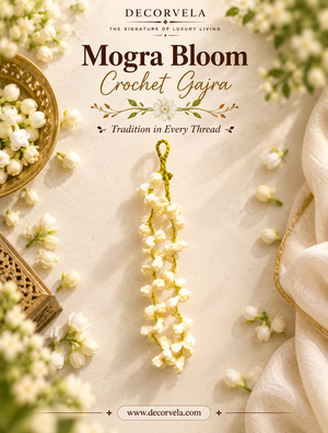 A decorative crochet garland made of small white blossoms hangs from a gold ring against a warm cream background, surrounded by floral props, fabric, and a bowl of white flowers. Large text at the top reads 'DECORVELA Mogra Bloom Crochet Gajra' with the website decorvela.