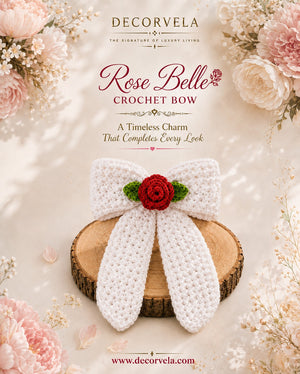 A white crocheted bow with a red rose and green leaves at the center, displayed on a round wood slice. The image is a styled product graphic with pink flowers in the corners and text reading 'DECORVELA Rose Belle Crochet Bow'.