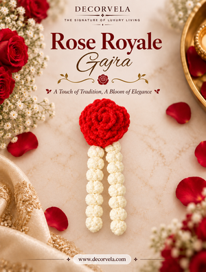 Decorvela Rose Royale Gajra, a traditional Indian embroidered flower garland, displayed on a beige marble surface with red roses, rose petals, and gold fabric accents. The garland features a vibrant red embroidered rose head with two white beaded stems.